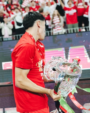 Vista lateral de un hombre en camiseta roja sosteniendo un trofeo de cristal roto, con público borroso y una pantalla digital detrás