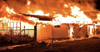 Una vivienda envuelta en grandes llamas durante la noche con una reja metálica en el frente y cables eléctricos en la parte superior