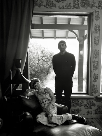 Fotografía en blanco y negro de una mujer sentada en un sillón y un hombre de pie junto a una ventana con vista a árboles