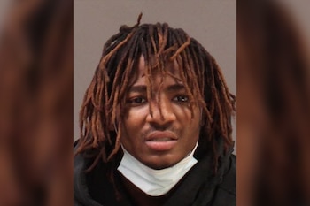Retrato frontal de un hombre joven con piel oscura y rastas castañas, mirando al frente. Lleva una mascarilla quirúrgica blanca en el cuello
