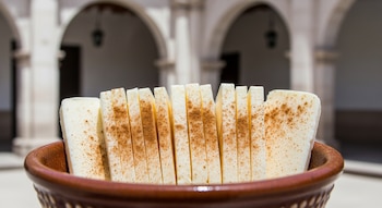 Queso helado en láminas dentro de un cuenco de cerámica, espolvoreado con canela. Al fondo, arcos de piedra sillar de un patio colonial.