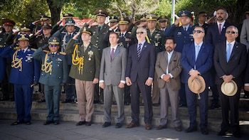 Un grupo de hombres, incluyendo militares en uniforme y civiles con traje, de pie en exteriores durante un evento; muchos de los militares saludan