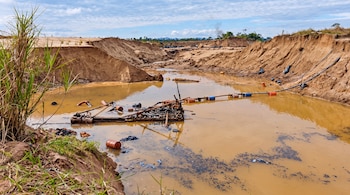 Poza de agua marrón entre montículos de arena cortados, con una plataforma de madera, barriles y tuberías.