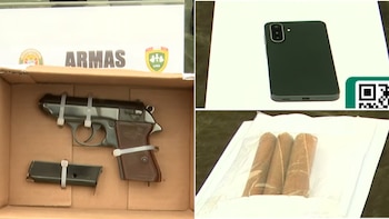 Una pistola negra con su cargador, un teléfono móvil oscuro y tres cartuchos de dinamita en una bolsa. Escudo y texto "ARMAS" en una placa blanca