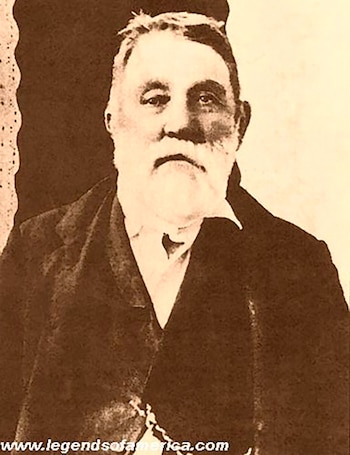 Fotografía sepia de un hombre mayor con barba y bigote blancos, vestido con un traje oscuro y camisa blanca, mirando directamente a la cámara