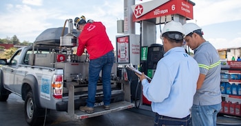 Tres hombres en una gasolinera Texaco. Uno trabaja en el equipo de una camioneta Toyota Tacoma gris, mientras dos con cascos miran un portapapeles