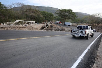 Una camioneta blanca circula por una carretera asfaltada cerca de la estructura metálica colapsada de un puente