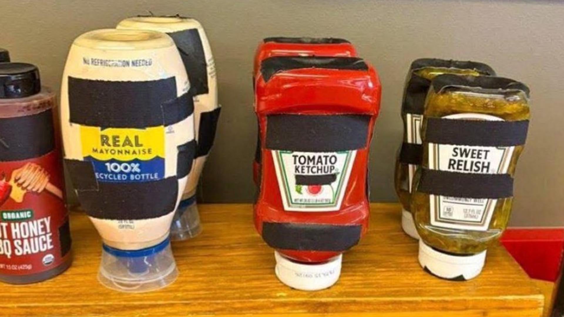 Varias botellas de condimentos, incluyendo mayonesa, kétchup, relish y salsa barbacoa, se exhiben en una superficie de madera con sus logotipos de marca cubiertos por cinta negra.