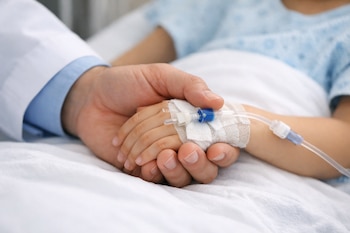 Mano de médico con bata blanca sosteniendo la mano de un niño con vía intravenosa en la muñeca y vendaje sobre una cama de hospital con sábanas blancas.