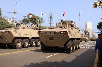 Varios vehículos militares blindados de color beige desfilan en una avenida. Soldados en los vehículos. Banderas de Perú y Corea del Sur. Edificios y público
