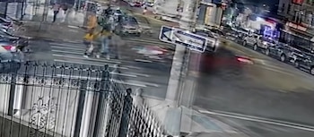 Vista aérea de una calle con vehículos estacionados y en movimiento, un paso de cebra, señales de tráfico, un semáforo y peatones