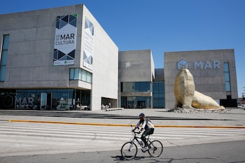 Edificio moderno de hormigón con letras MAR, escultura dorada de lobo marino, persona en bicicleta, cruce peatonal y cielo azul despejado
