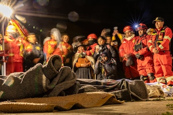 Grupo de personas con uniformes rojos de emergencia y civiles en escena nocturna con luces; varias mantas y un rollo de tela en el suelo