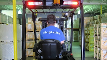 Vista trasera de un hombre operando un montacargas en un almacén industrial con estanterías llenas de mercancías embaladas y cajas