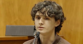 Primer plano de un joven con cabello rizado y tez clara, vistiendo una camisa marrón, hablando a un micrófono con un fondo de pared de madera