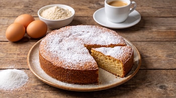 Un pastel espolvoreado con azúcar, una porción cortada, tres huevos, un cuenco con harina y una taza de café sobre una mesa de madera.