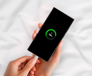 Dos manos sostienen un teléfono móvil negro con la pantalla encendida mostrando el estado de carga al 100 por ciento, conectado a un cable blanco