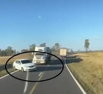 Un automóvil blanco detenido en la banquina de una carretera de dos carriles, al lado de un camión blanco y otro camión en la distancia