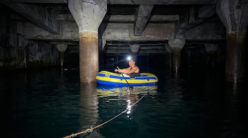 Joven en balsa inflable amarilla y azul rema en agua oscura bajo una estructura de hormigón con grandes pilares. Lleva linterna frontal encendida.