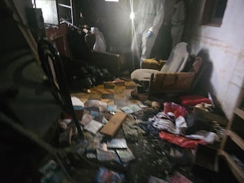 En una habitación oscura y desordenada, personal forense con trajes protectores blancos ilumina con linternas mientras investigan una escena del crimen