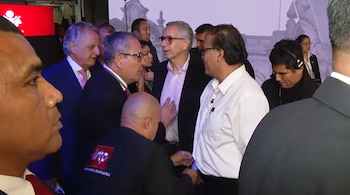 Un grupo de hombres en trajes y camisas conversando en un evento con pantallas al fondo; uno lleva una camiseta negra con el logo de IRTP en la espalda.