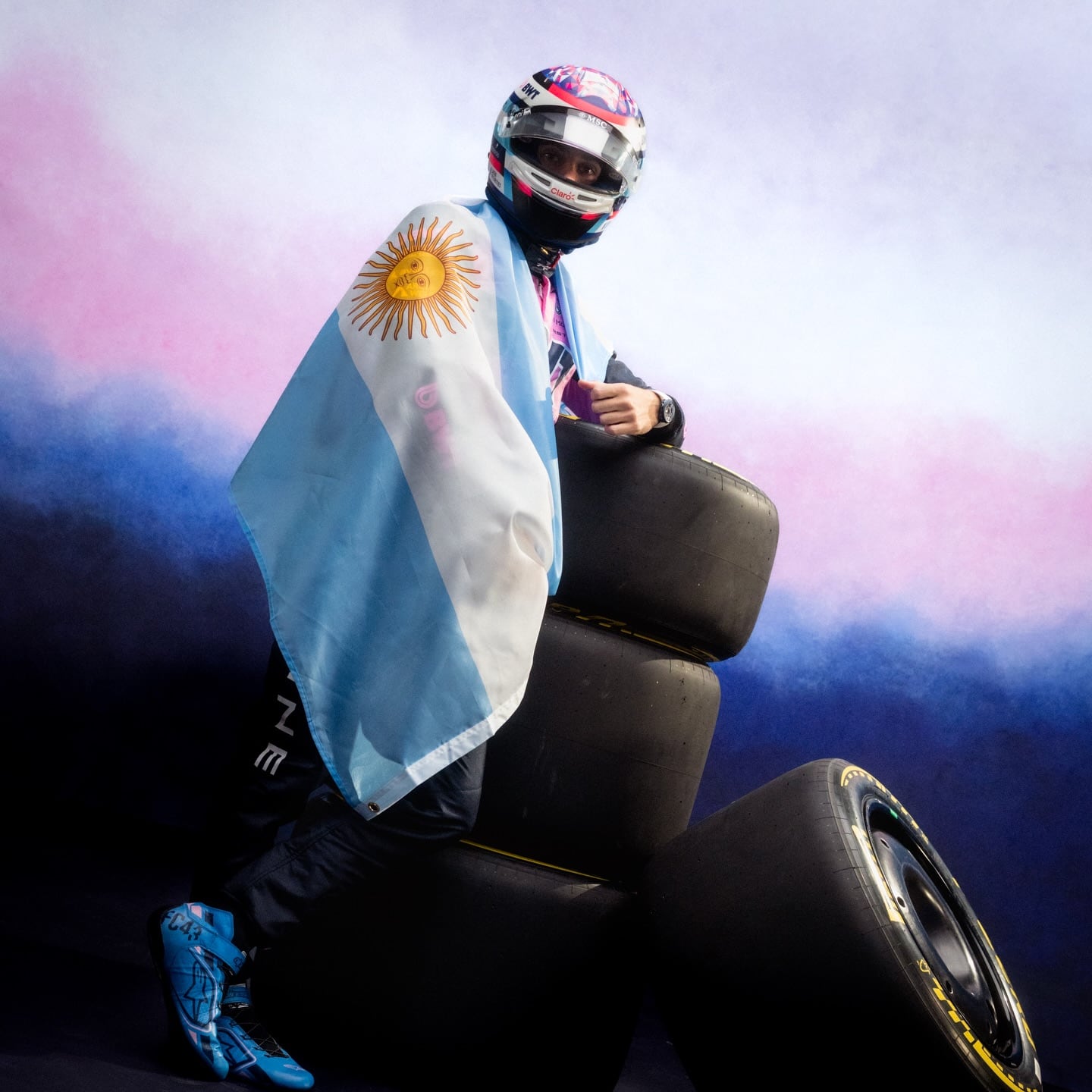 El piloto argentino Franco Colapinto posa con orgullo envuelto en la bandera nacional, apoyado sobre neumáticos de carrera, en un estudio con fondo de colores vibrantes.