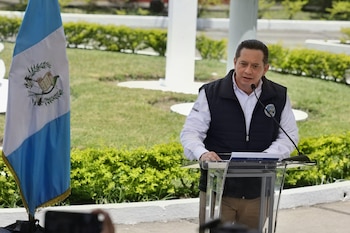 Un hombre en un podio transparente, con camisa blanca y chaleco oscuro con el logo de INSIVUMEH, habla. A su izquierda, se ve la bandera de Guatemala. Fondo exterior verde