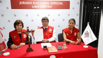 Tres autoridades con chalecos rojos del Jurado Nacional de Elecciones están sentadas ante una mesa con un crucifijo y un documento