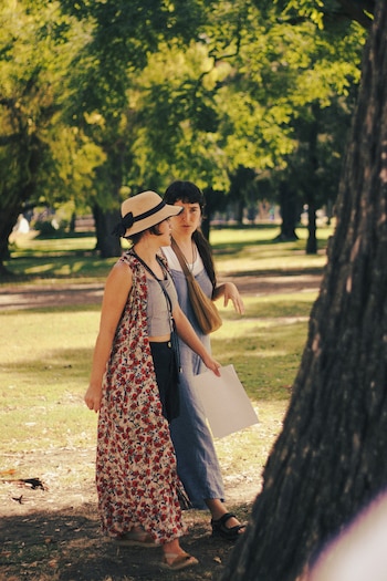 Dos mujeres caminan en un parque. Una lleva sombrero de paja y vestido floral, la otra viste un peto y sujeta papeles blancos. Fondo de árboles verdes y pasto