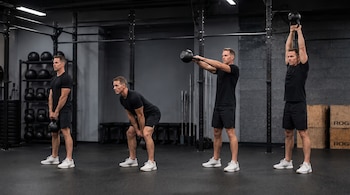 Un hombre vestido con camiseta y pantalones cortos negros realiza un swing con pesa rusa en cuatro etapas. Hay pesas y equipamiento de gimnasio al fondo.