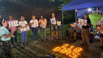 Varias personas de pie sostienen carteles con retratos alrededor de decenas de pequeñas velas encendidas en el suelo durante la noche