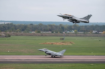 Dos aviones de combate militares grises en una pista de aterrizaje con zonas de césped y árboles al fondo