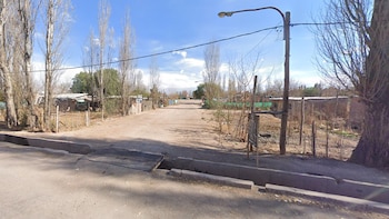 Una calle de tierra flanqueada por árboles altos, casas precarias, un poste de luz con tendido eléctrico y una zanja con puente metálico