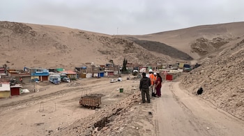 Personas en un camino de tierra, una con casco y chaleco, frente a viviendas dispersas, vehículos y colinas de arena.