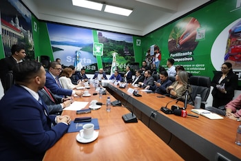 Mesa larga de conferencia, personas sentadas y de pie, micrófonos, tazas, botellas de agua, papeles. Paredes verdes con gráficos de paisajes, logo UNE y texto