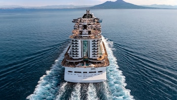 Vista aérea trasera del crucero blanco MSC Seaview navegando en el mar azul, dejando una estela. Se aprecian terrazas con personas y montañas al fondo