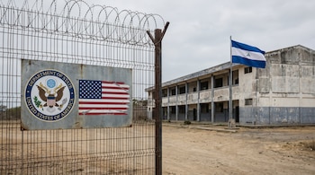 Valla metálica con alambre de púas, cartel de Departamento de Estado de EE. UU. y bandera estadounidense. Al fondo, bandera de Nicaragua y edificio gris deteriorado.