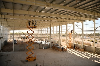Interior de un hangar en construcción con vigas metálicas en el techo, piso de cemento, cajas y operarios sobre dos plataformas elevadoras amarillas