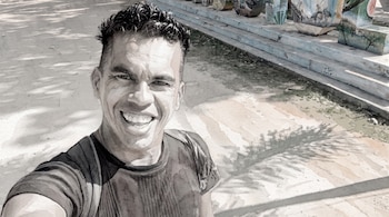 Primer plano de un hombre sonriente mirando a cámara, con pelo corto oscuro y camiseta. El fondo muestra un suelo y estructuras pintadas con efecto acuarela.