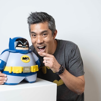 Marcelo Wong sonríe y señala una escultura de Batman de formas redondeadas apoyada sobre una estructura blanca