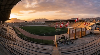 Vista panorámica de un estadio de fútbol vacío con pista de atletismo, tribunas, banderas de Perú y flores en el ingreso al atardecer.