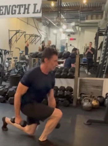 Hombre realiza estocadas con mancuernas en un gimnasio con varias pesas y equipamiento de ejercicio. Varias personas al fondo