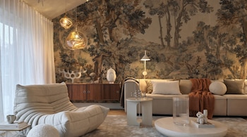 Vista frontal de un salón moderno con un tapiz de bosque en la pared. Incluye un sofá modular, un sillón lounge, mesas auxiliares, lámparas y cortinas blancas.