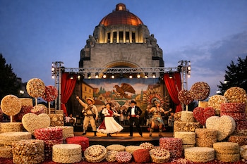 Monumento a la Revolución detrás de un escenario de teatro con telones rojos, un fondo pintado y cuatro actores vestidos con trajes, con dulces de amaranto en primer plano.