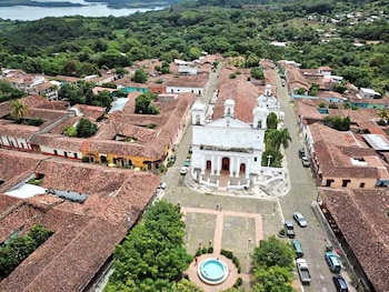 Vista aérea de un pueblo con una iglesia blanca grande, edificios de techos rojos, una plaza con fuente, árboles verdes y un lago rodeado de montañas