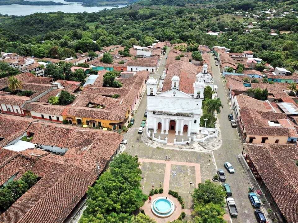 Una vista panorámica muestra la arquitectura colonial de un pueblo del oriente de El Salvador, destacando su iglesia blanca, tejados de barro y una plaza central, enmarcado por exuberante vegetación y un cuerpo de agua al fondo.