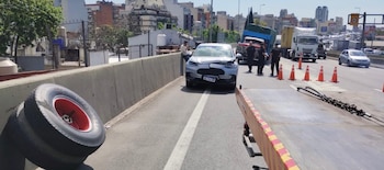 Un vehículo gris con el capó abollado, dos neumáticos de camión apoyados en el muro, conos naranjas y policías en una autopista