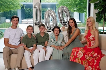 Una familia de seis personas posa sentada en un sofá blanco delante de tres globos plateados que forman el número cien