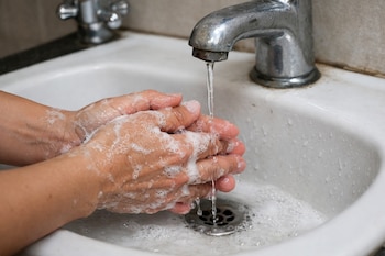 Manos con jabón se enjuagan bajo un grifo con un chorro de agua delgado en un lavabo blanco con desagüe.