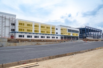 Vista exterior de un edificio comercial blanco con detalles amarillos y azules en construcción. Al lado derecho, una gran estructura metálica azul. En primer plano, una carretera y terreno con obreros